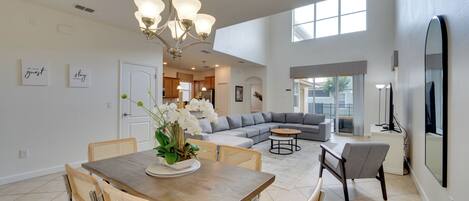 A spacious living room with a large gray sectional sofa, a dining area with a wooden table and wicker chairs, and a kitchen in the background.