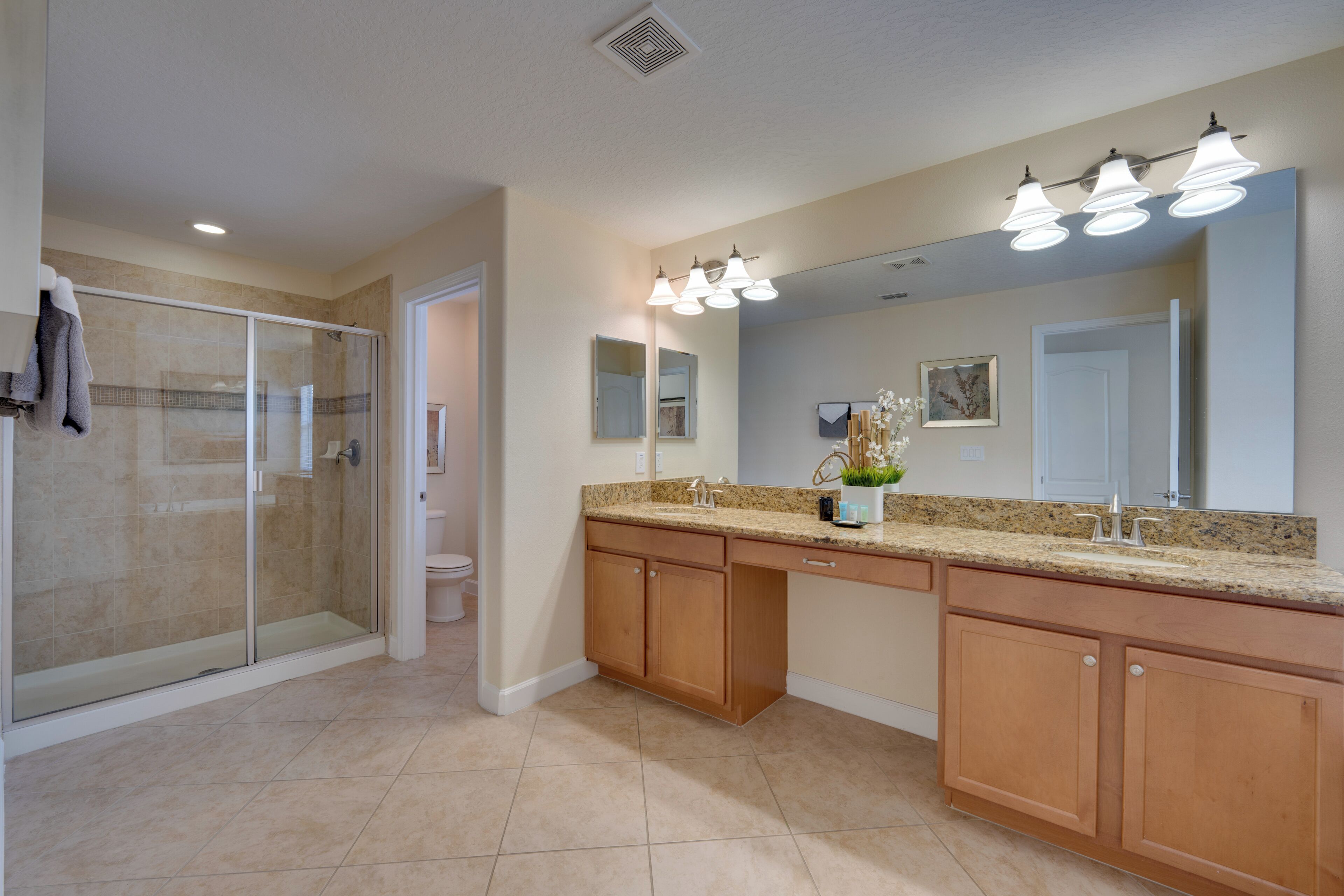 A modern bathroom with a glass shower enclosure, a large vanity with a granite countertop, and a wall-mounted mirror with three lights.