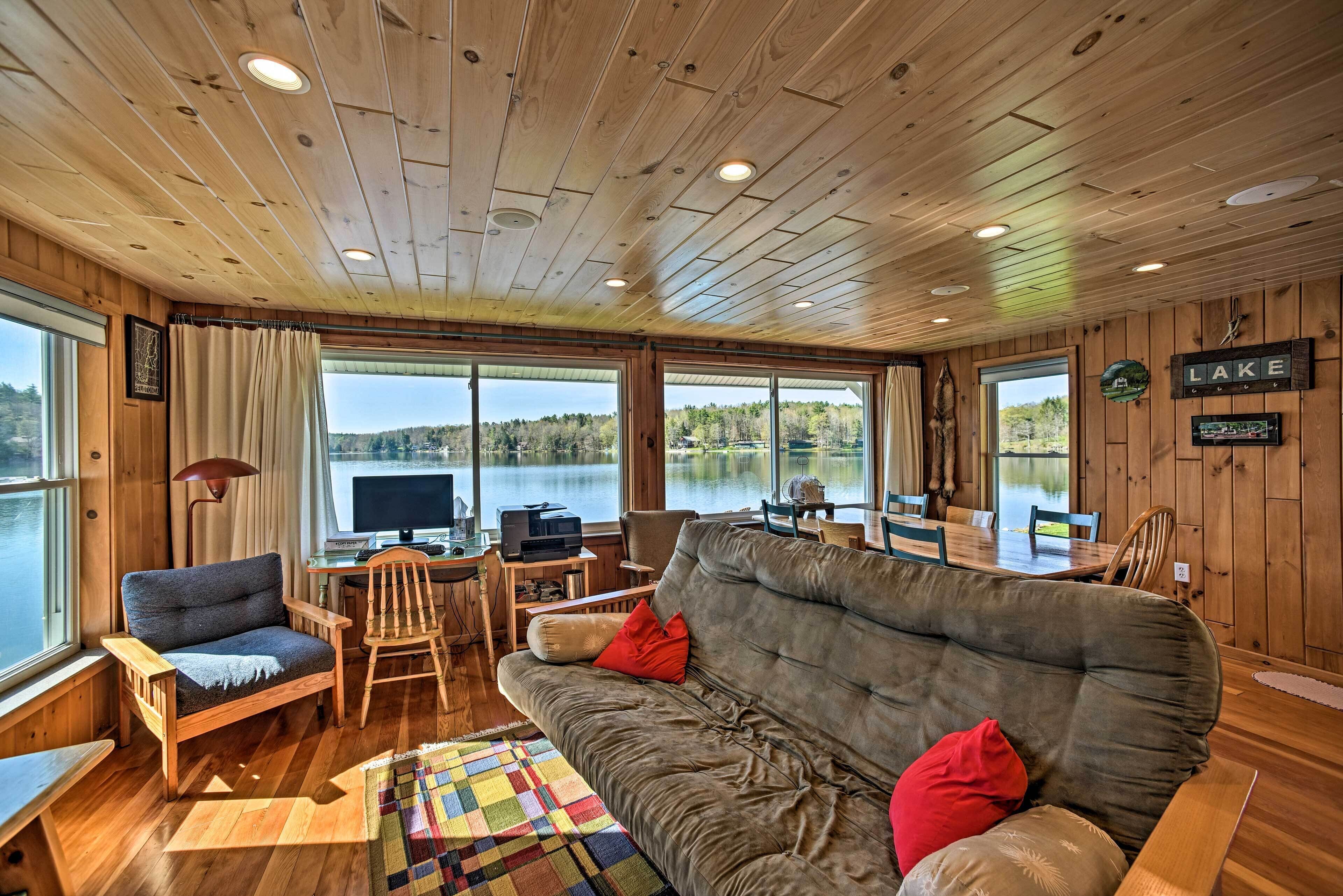 A cabin with a wooden interior, a large window overlooking a lake, a sofa, a desk with a computer, and a dining area.