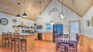 Une cuisine spacieuse avec un plancher en bois, un îlot central, des tabourets de bar, un éclairage suspendu et un panneau décoratif au mur avec l’inscription « rassemblement ».