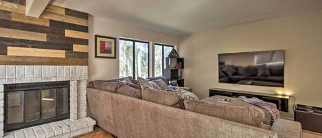 A living room with a sectional sofa, a fireplace, a flat-screen TV, and large windows.