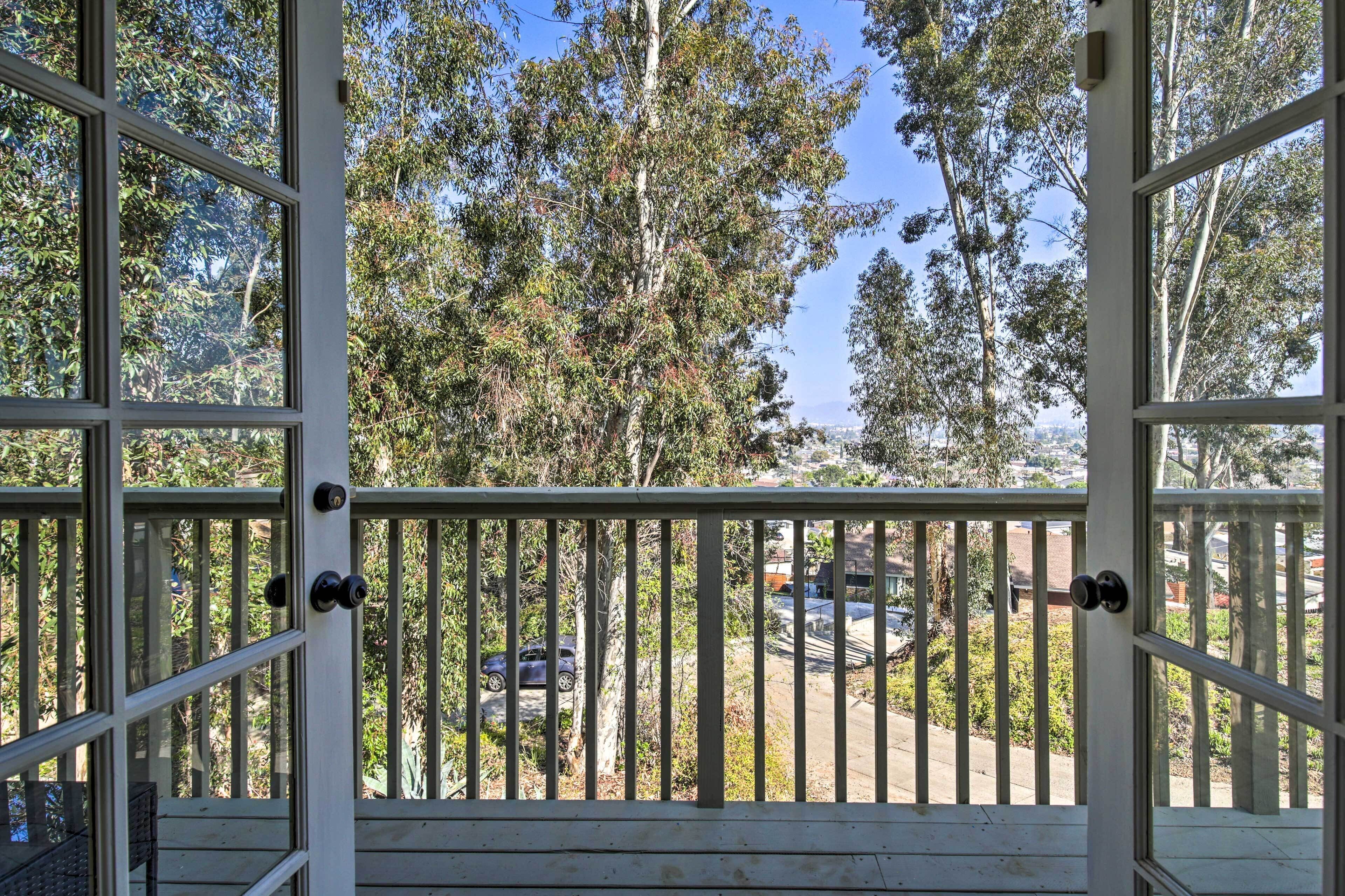 A view from a window with a white frame and a wooden railing, overlooking a residential area with trees and parked cars.
