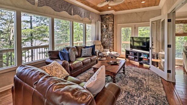 A spacious living room with leather sofas, a stone accent wall, a large TV, and a balcony with a view of trees.