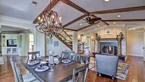 A spacious dining area with a wooden table set for six, surrounded by upholstered chairs. A chandelier hangs above the table, and a fireplace is visible in the background.