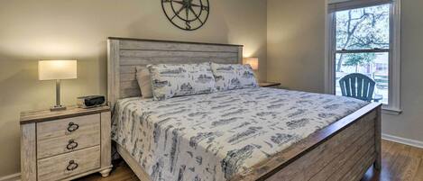 A bedroom with a wooden bed, a nightstand with drawers, a compass wall decoration, and a window with a view of the outside.