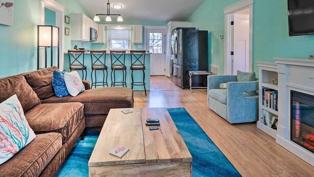 A living room with a brown sectional sofa, a wooden coffee table, and a blue rug. There is a kitchen area with white cabinets and a dining set. A fireplace is visible on the right side.