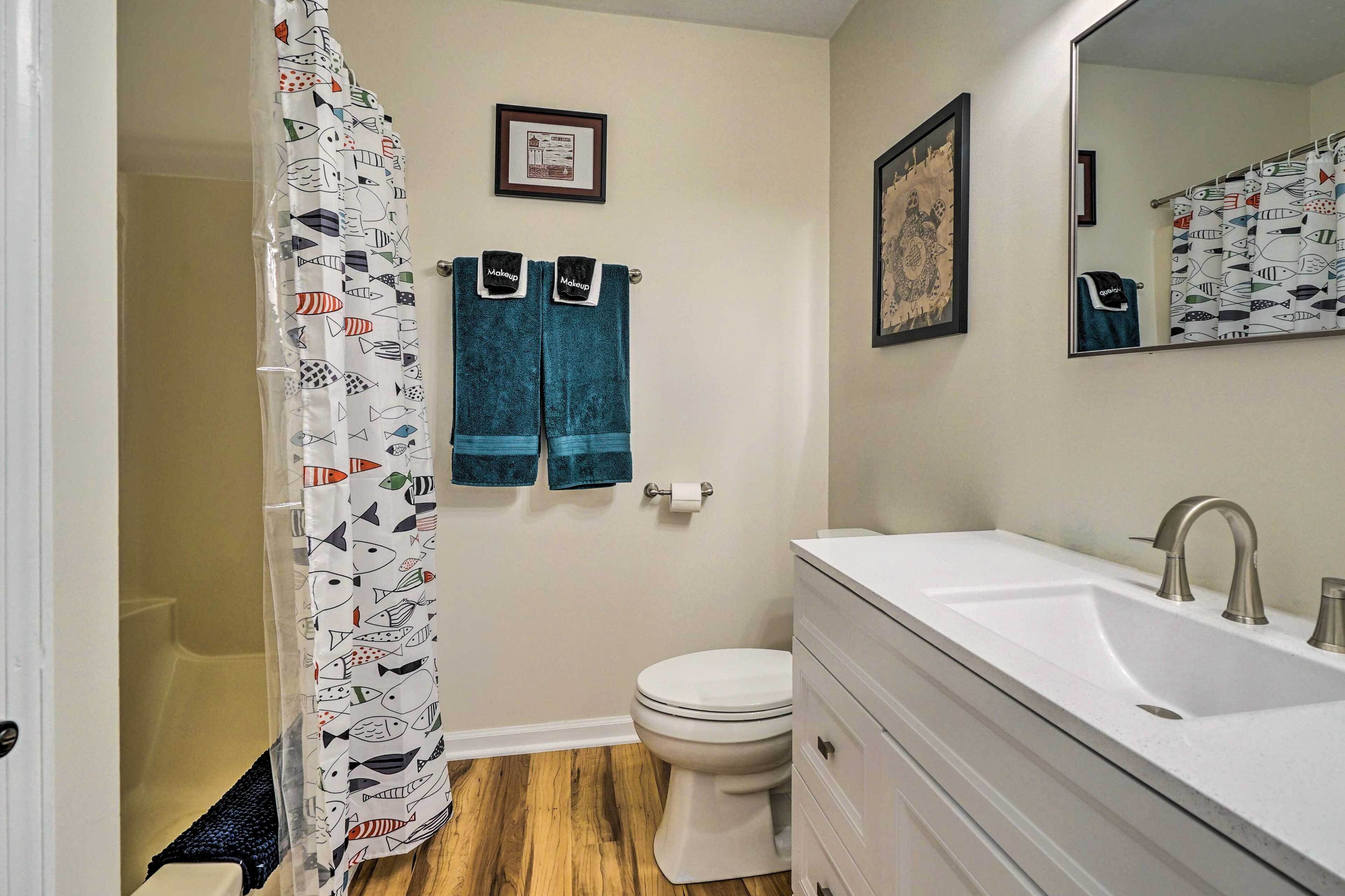Une salle de bain avec un lavabo blanc, des toilettes et un rideau de douche orné d’un motif de poissons. Deux serviettes sont accrochées au mur et un tableau encadré est placé au-dessus des toilettes.
