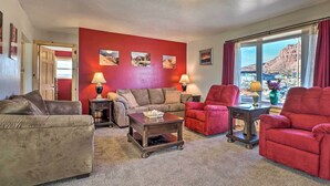 A living room with a red wall, a sofa, two armchairs, a coffee table, and a view of a mountainous landscape through large windows. - Rural Base Camp for Grand Canyon, Zion & Bryce NP! (Kanab)