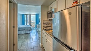 A modern kitchen with stainless steel appliances, a microwave, and a refrigerator. There is a balcony with blue curtains and a bed in the adjacent room. - Steps to Shore: Beachfront Studio w/ Pool Access! (Myrtle Beach)