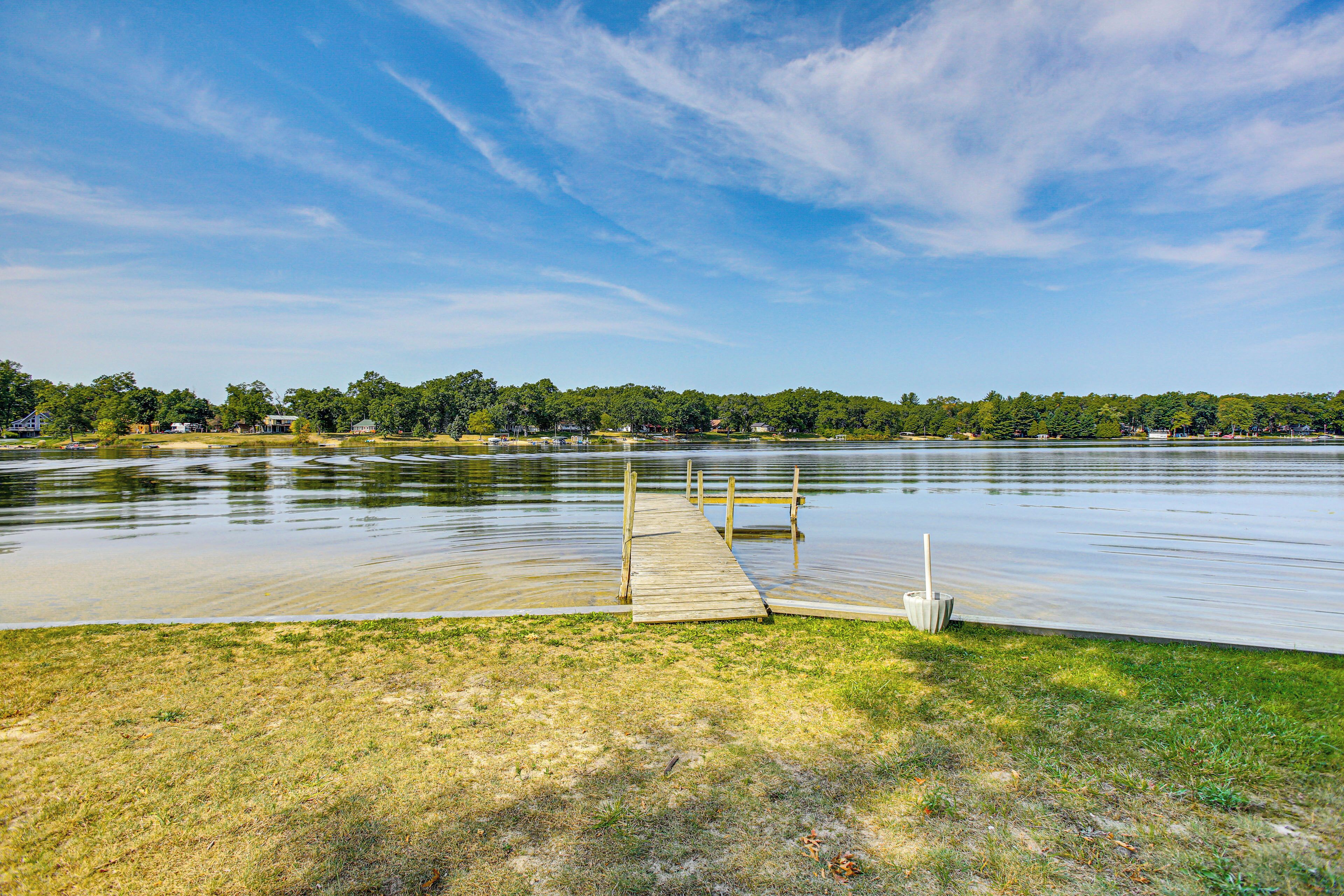 A wooden dock extending into a calm lake with a grassy shore and trees in the background.