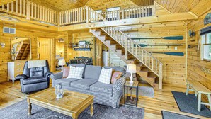 A spacious cabin interior with a checkered sofa, a leather chair, a wooden dining table, and a staircase leading to an upper level. - Peaceful Long Lake Cottage w/ Deck, Dock & Kayaks! (Branch)