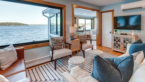 A modern living room with a large window overlooking a body of water, a television mounted on the wall, and a cozy seating area with patterned cushions. - Winnipesaukee Lakefront Home With Dock & Views! (Alton Bay)