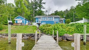 A wooden dock leading to a blue house with a porch and outdoor furniture. - Winnipesaukee Lakefront Home With Dock & Views! (Alton Bay)