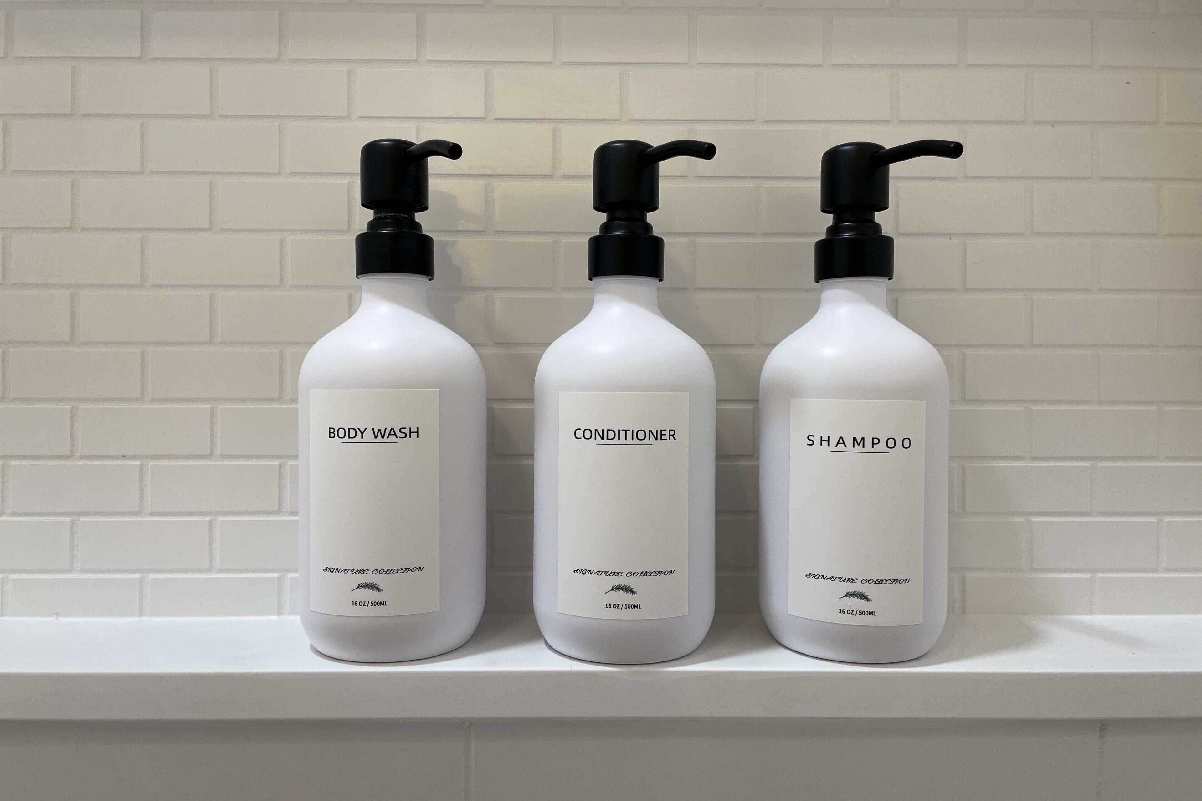 Three bottles of body wash, conditioner, and shampoo on a white shelf against a white brick wall.