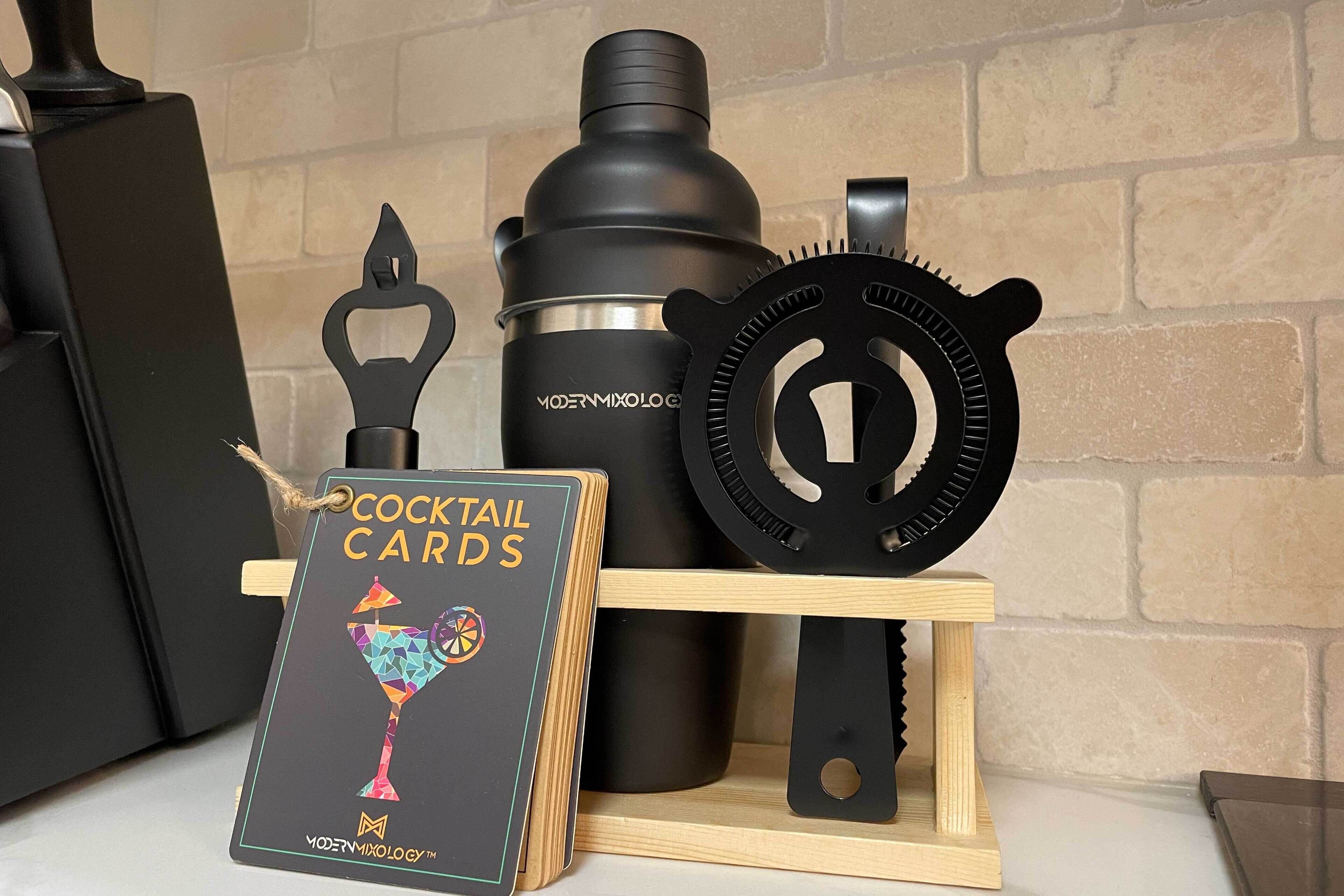 A cocktail card, a black shaker, and a cocktail shaker strainer on a wooden stand.