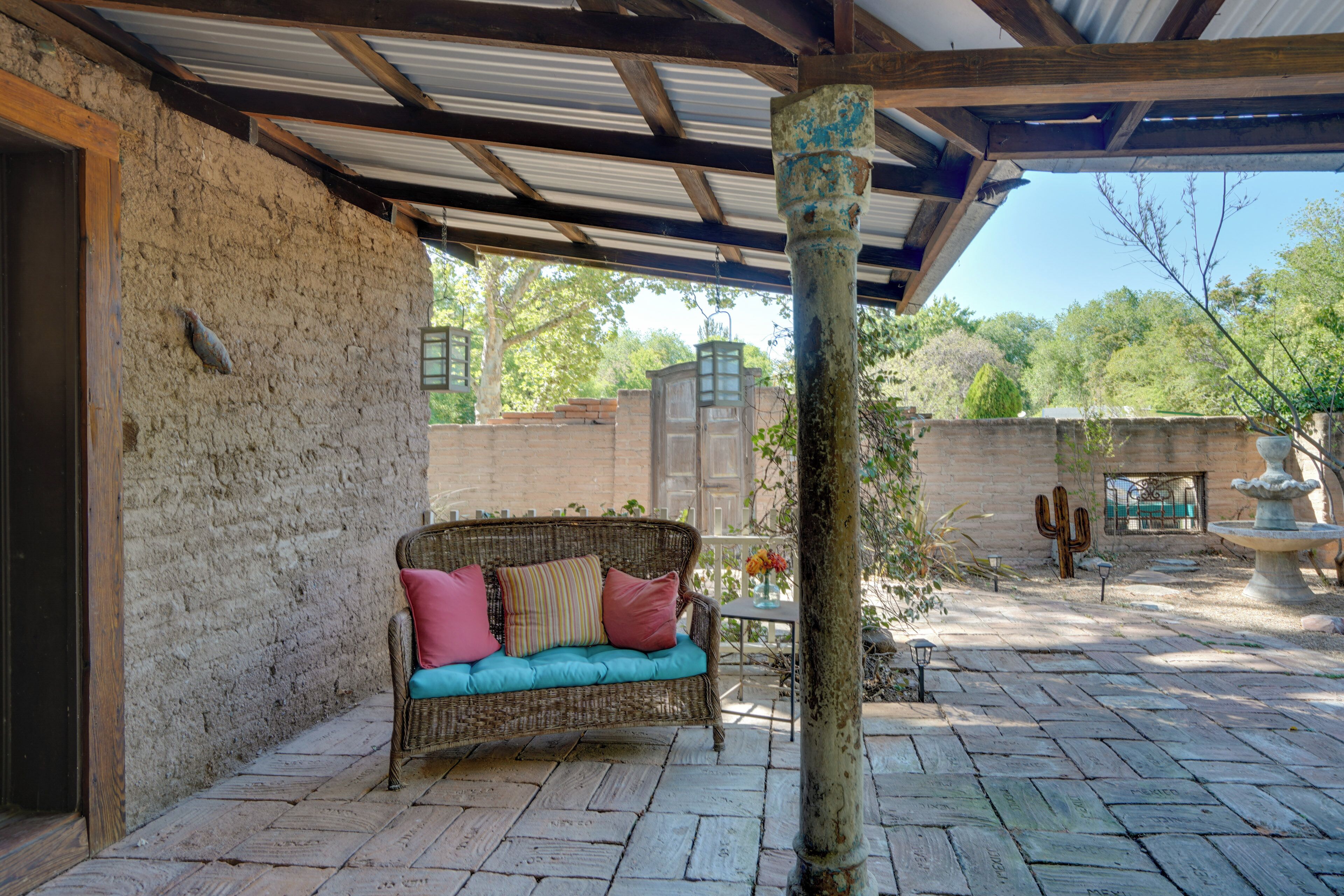 A covered patio with a wicker sofa, a stone pillar, and a cactus in the background.