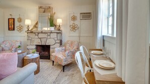 Un salon chaleureux avec une cheminée en pierre, des fauteuils à motifs, une table à manger dressée pour quatre personnes et une grande fenêtre laissant entrer la lumière naturelle.