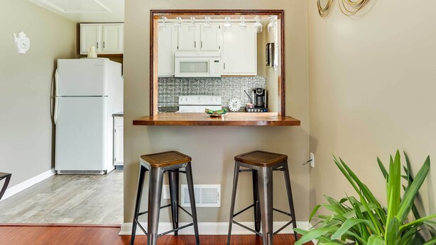A compact kitchen with white cabinets, a microwave, and a refrigerator. There are two stools and a potted plant. - Walkable Apt Near Kentucky Derby! (Louisville)