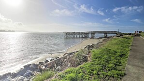 Une jetée s’avançant dans des eaux calmes, une plage de sable et une zone herbeuse avec un chemin en béton.