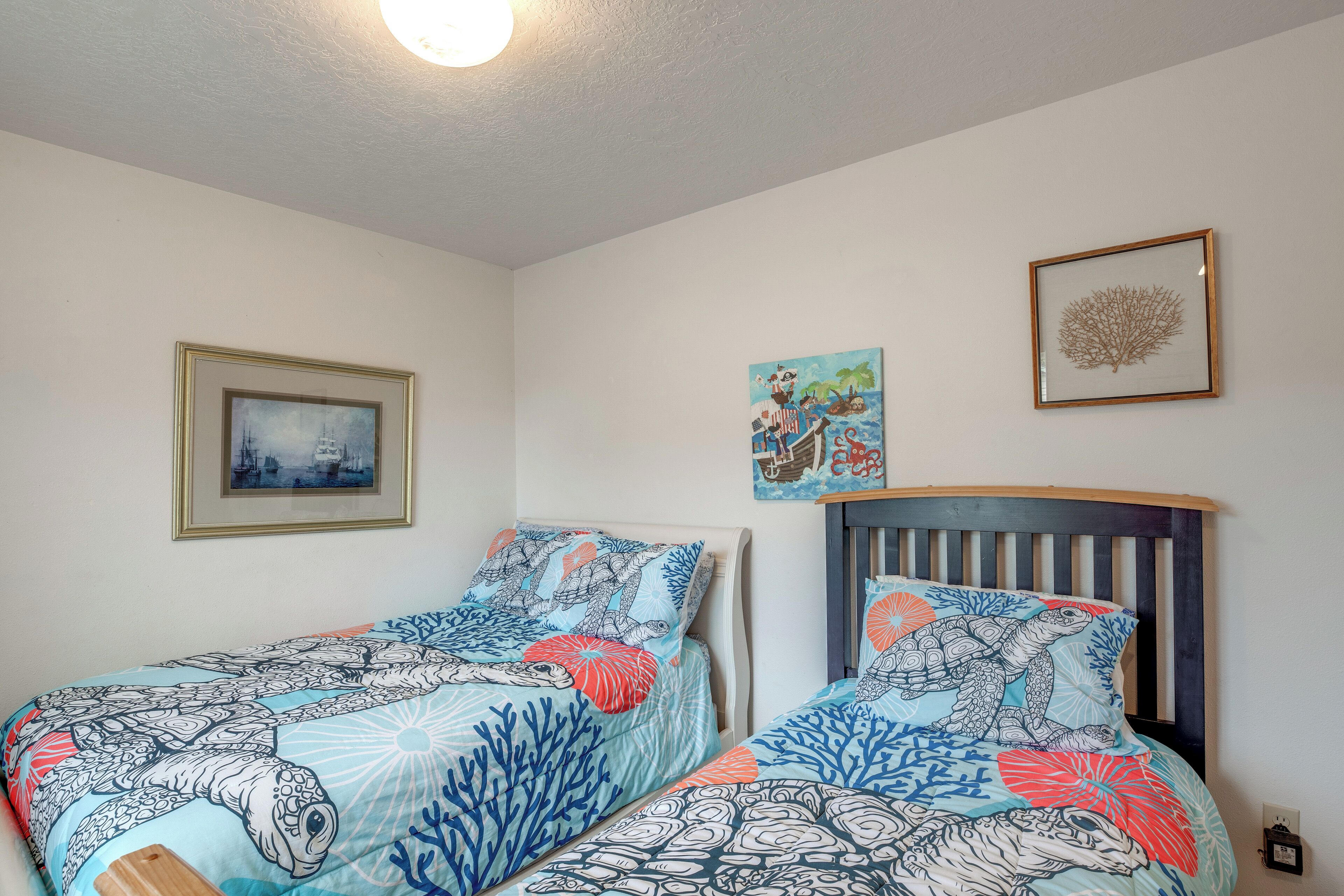 Ein Schlafzimmer mit zwei Betten, jedes mit einer individuellen Bettdecke im Meeresschildkröten-Design. An den Wänden hängen Bilderrahmen, eines zeigt ein Schiff und das andere einen Baum.