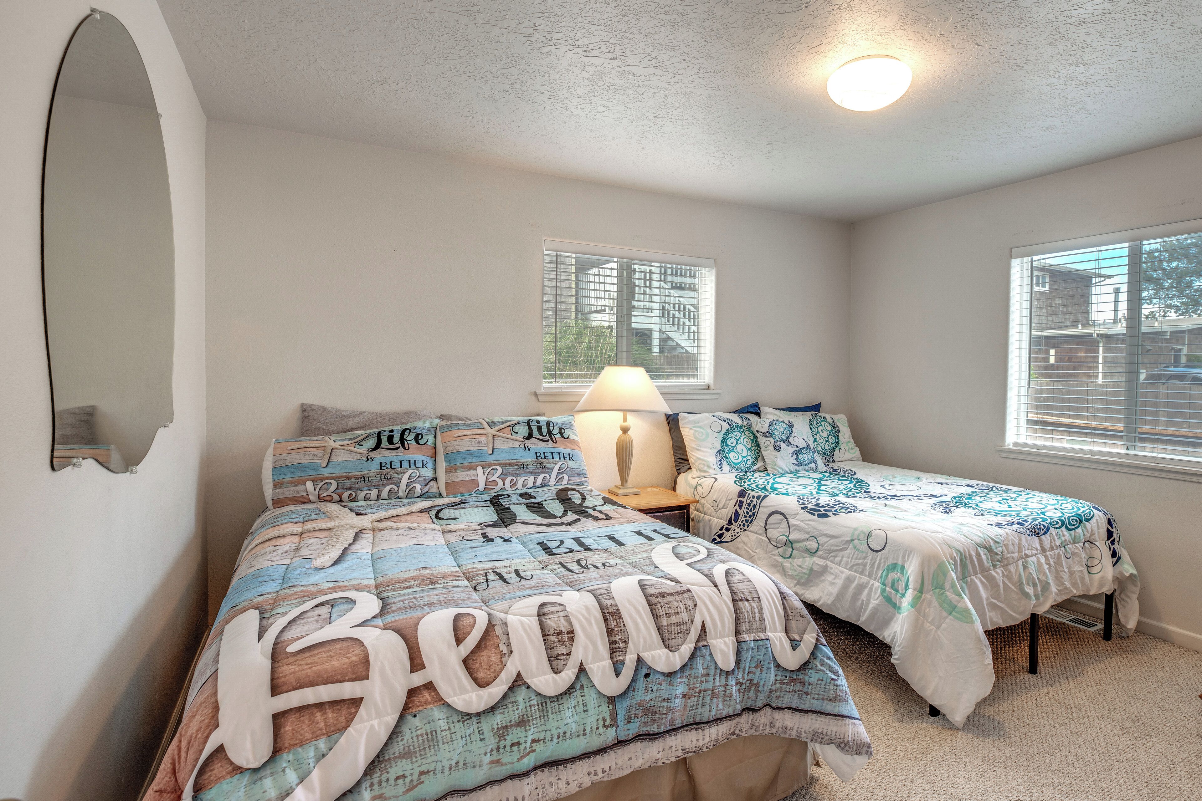 Une chambre avec deux lits, chacun recouvert d’une couvre-lit sur le thème de la plage. Il y a une fenêtre avec des stores, une lampe sur la table de chevet et un miroir au mur.