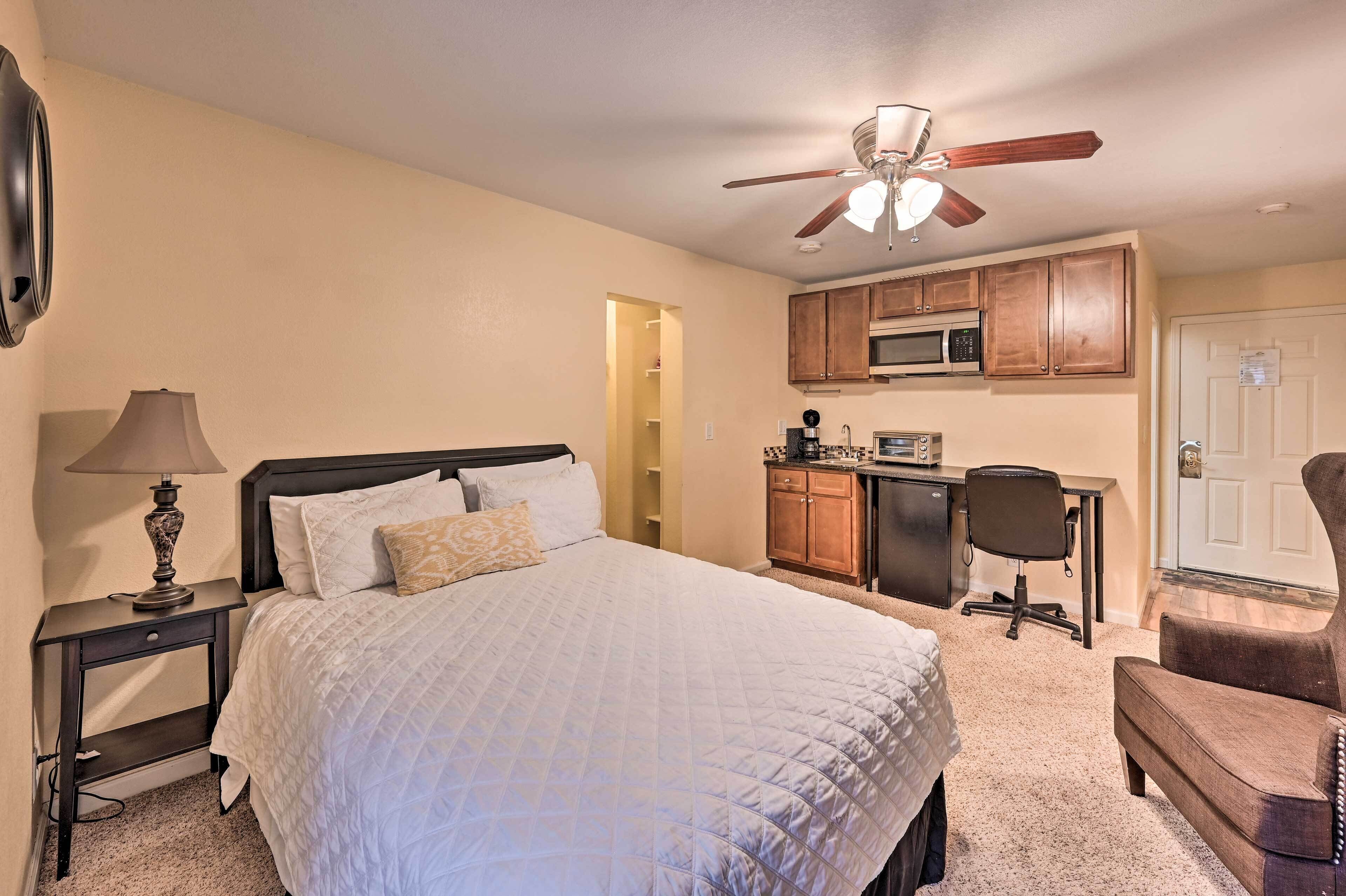 A bedroom with a bed, bedside table, lamp, ceiling fan, and a kitchenette with a microwave and cabinets.