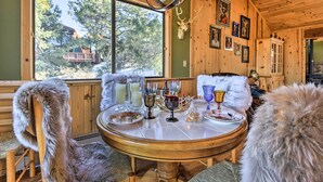 Un intérieur en bois avec une table à manger dressée, une grande fenêtre donnant sur un paysage arboré et un crâne de cerf accroché au mur.