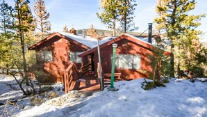 Une cabane rouge, dont le toit est recouvert de neige, entourée d’arbres et sous un ciel dégagé.