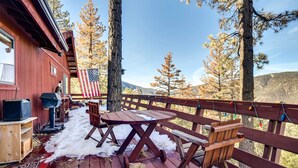 Une terrasse en bois avec une table et des chaises, un drapeau américain, et une vue sur les arbres et les montagnes.