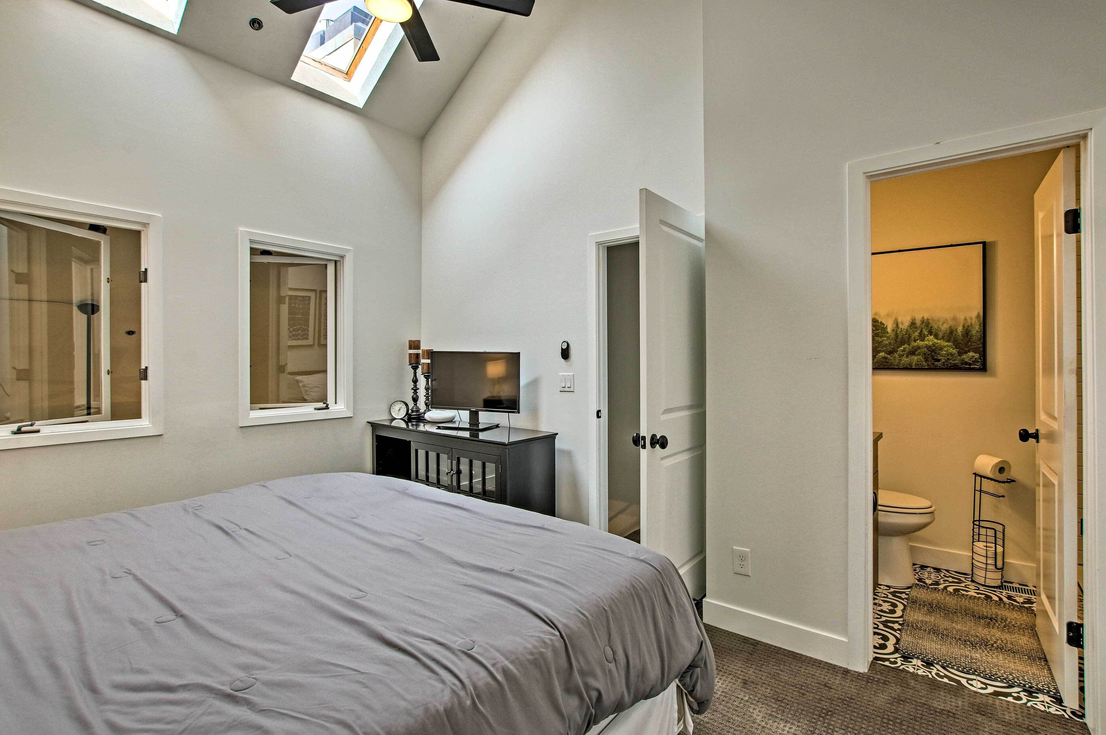 A bedroom with a bed, a desk with a computer, a window, and a door leading to a bathroom.