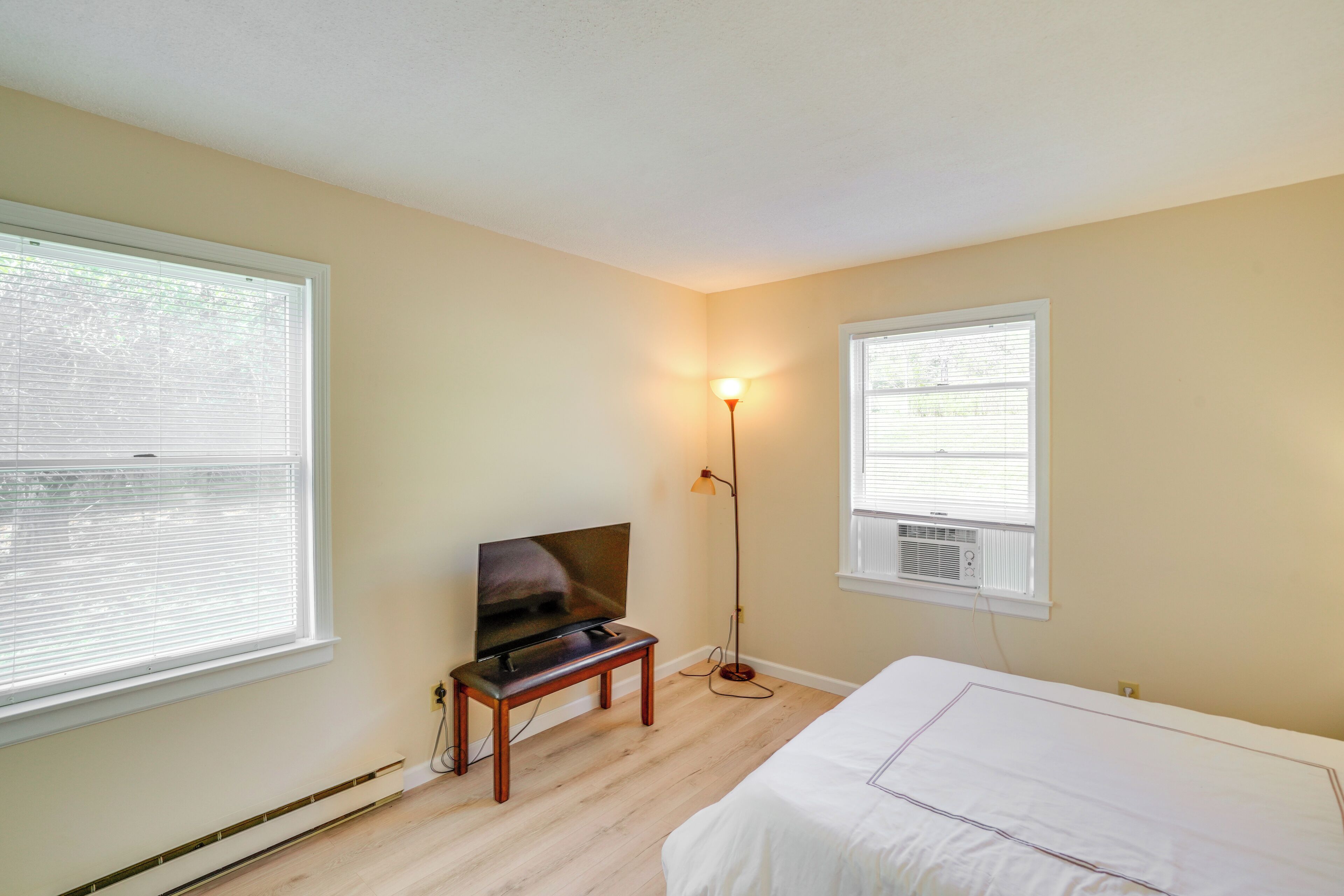 A bedroom with a bed, a TV on a stand, a floor lamp, and two windows with blinds.