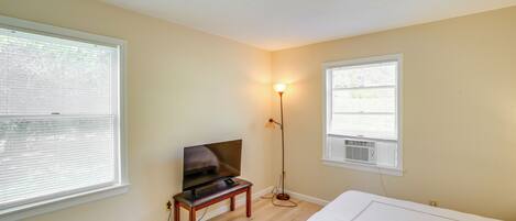 A bedroom with a bed, a TV on a stand, a floor lamp, and two windows with blinds.