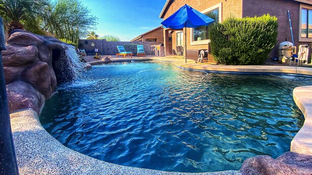 A swimming pool with a waterfall feature, surrounded by lounge chairs and a building with a tiled roof. - Fire Pit, Covered Patio: Stylish Goodyear Refuge (Goodyear)