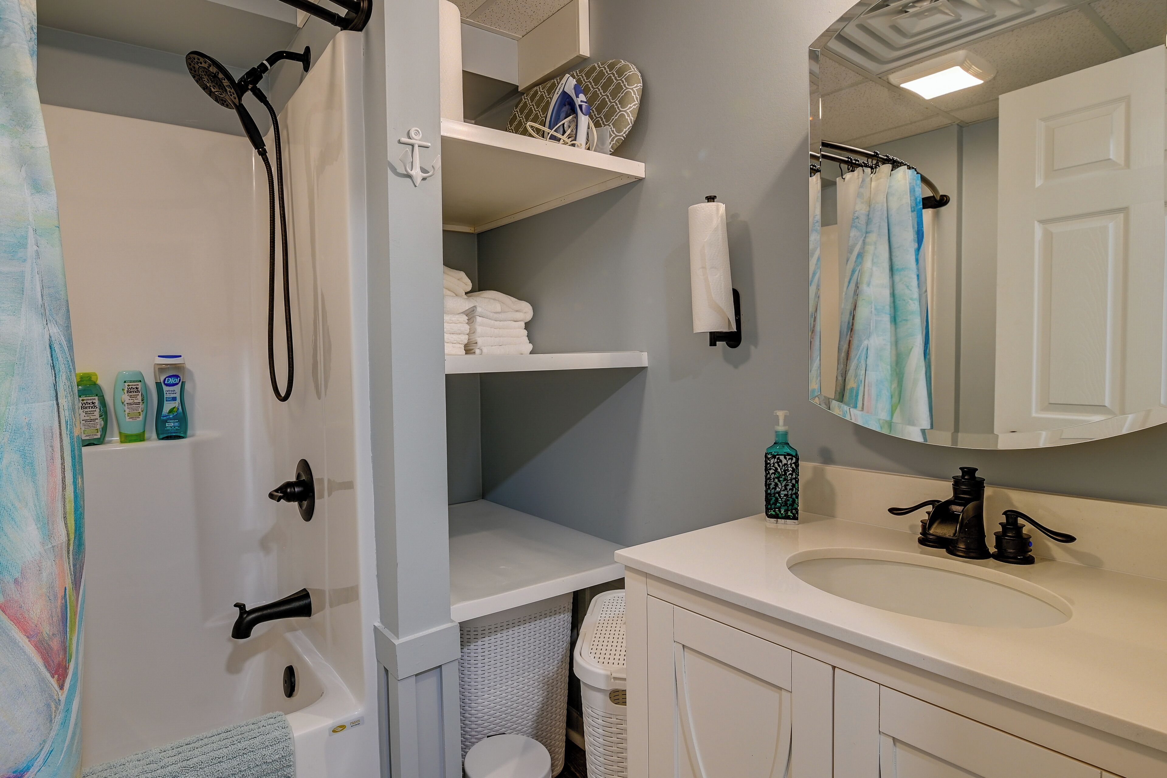 A bathroom with a white bathtub, a sink with a mirror above it, and shelves above the bathtub holding towels and toiletries.