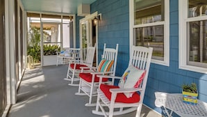 A covered porch with white rocking chairs and red cushions, a small table with a plant, and a blue exterior wall. - 'below Deck' Beach House: Walk to the Ocean! (Murrells Inlet)