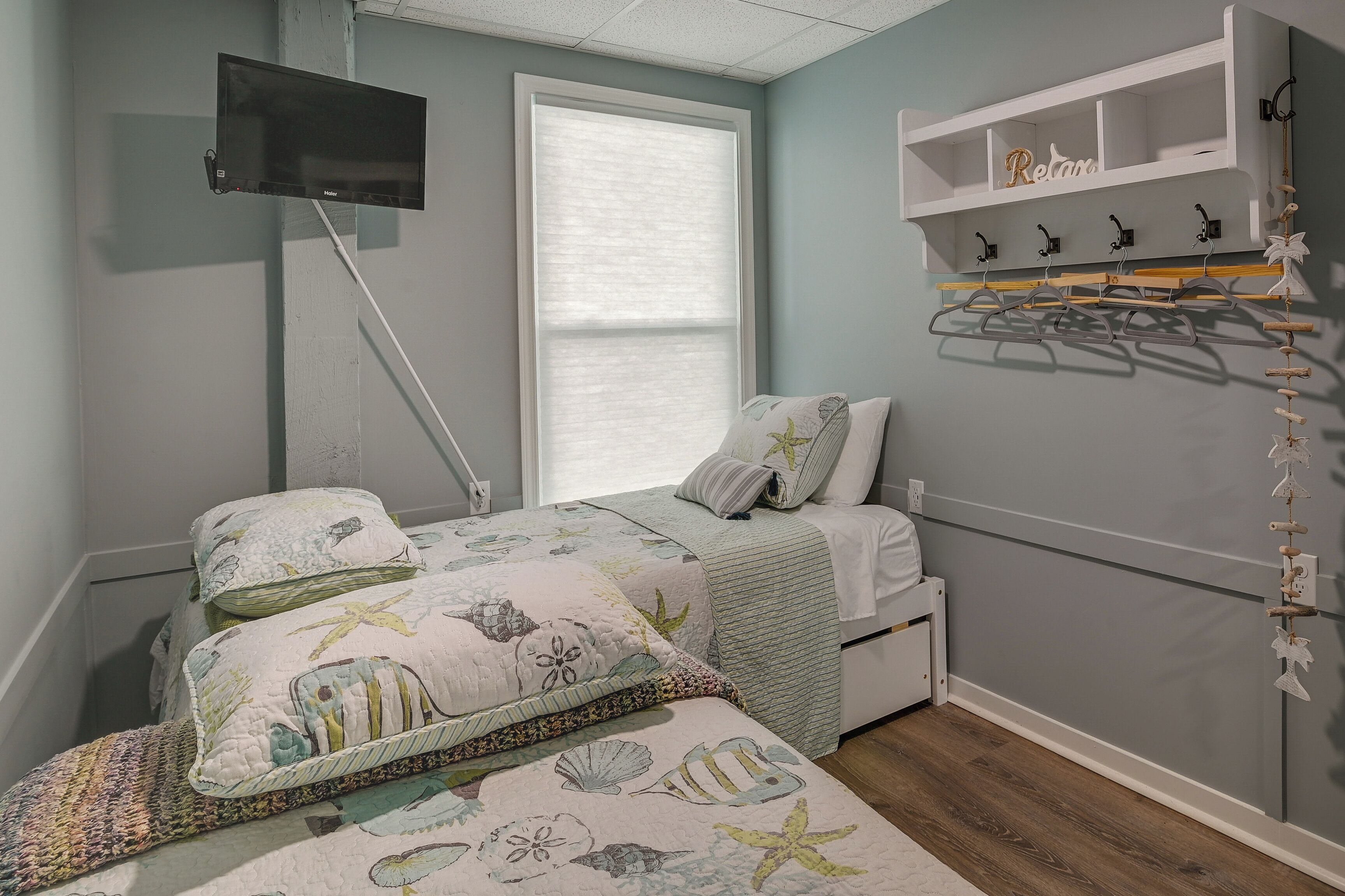 Une chambre à coucher comprenant un lit, une table de chevet, un téléviseur fixé au mur, une fenêtre avec des stores et une étagère sur laquelle sont suspendus divers objets.