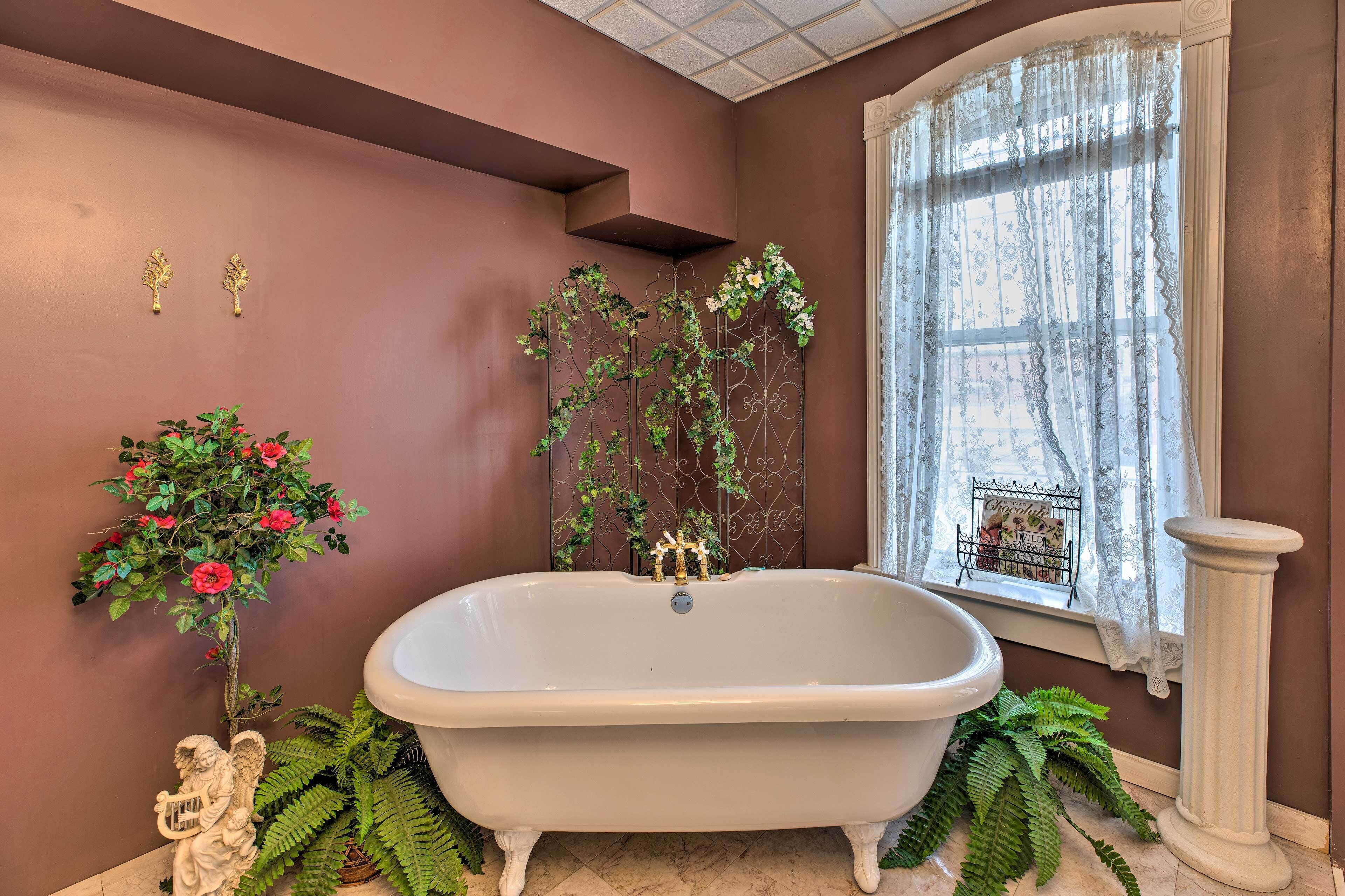 A bathroom with a freestanding bathtub, a window with lace curtains, and decorative plants.
