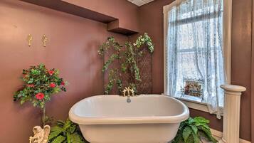 A bathroom with a freestanding bathtub, a window with lace curtains, and decorative plants.