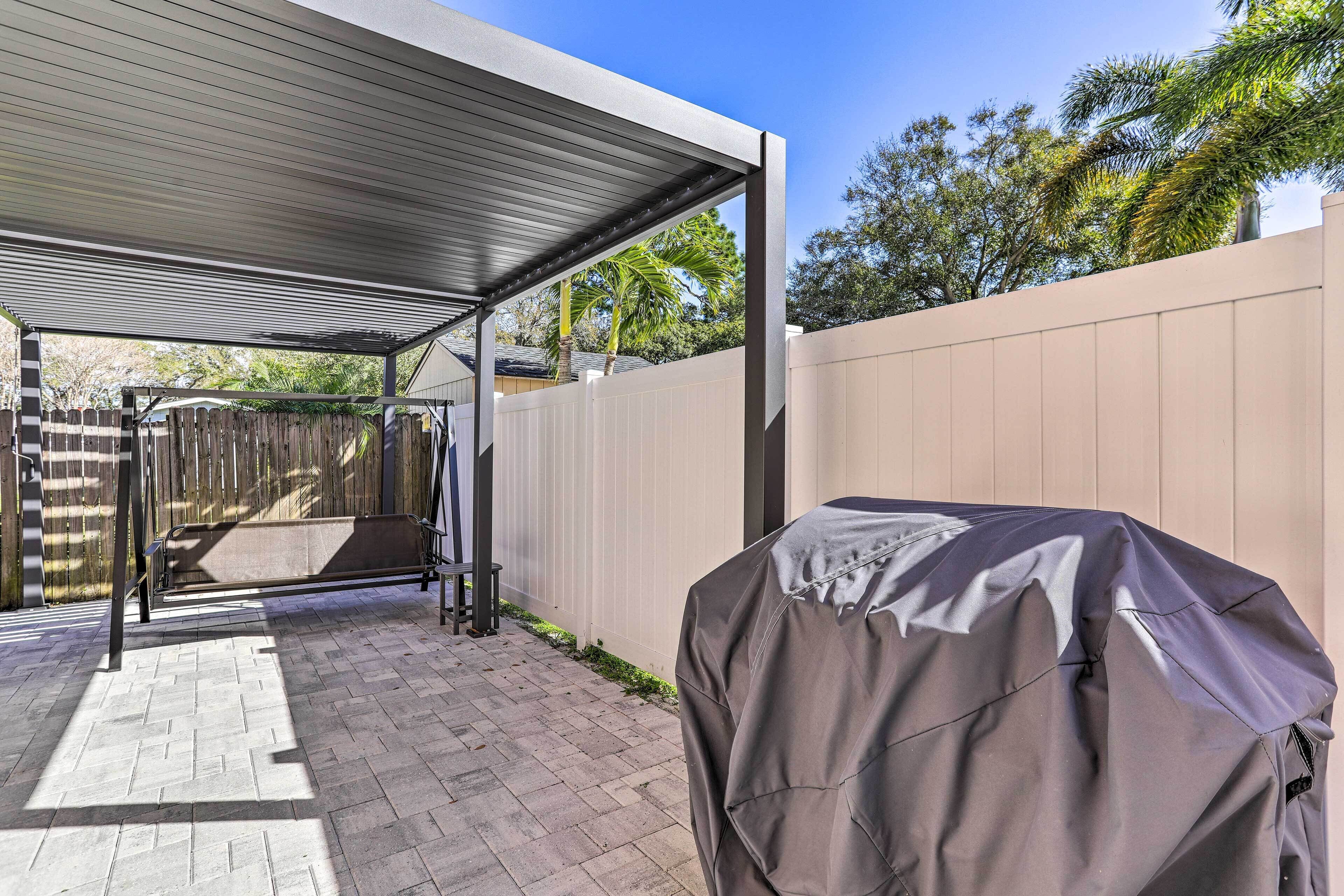 A covered patio area with a barbecue grill, a wooden fence, and a paved walkway.