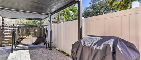 A covered patio area with a barbecue grill, a wooden fence, and a paved walkway.