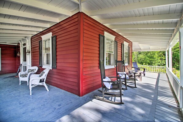 Une maison en bois rouge avec un porche, des chaises en osier blanches et un fauteuil à bascule.