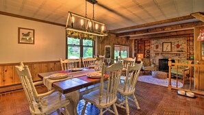 A dining area with a wooden table and chairs, a chandelier, and a fireplace in the background. - Vintage Schroon Lake Farmhouse w/ Private Beach! (Schroon Lake)