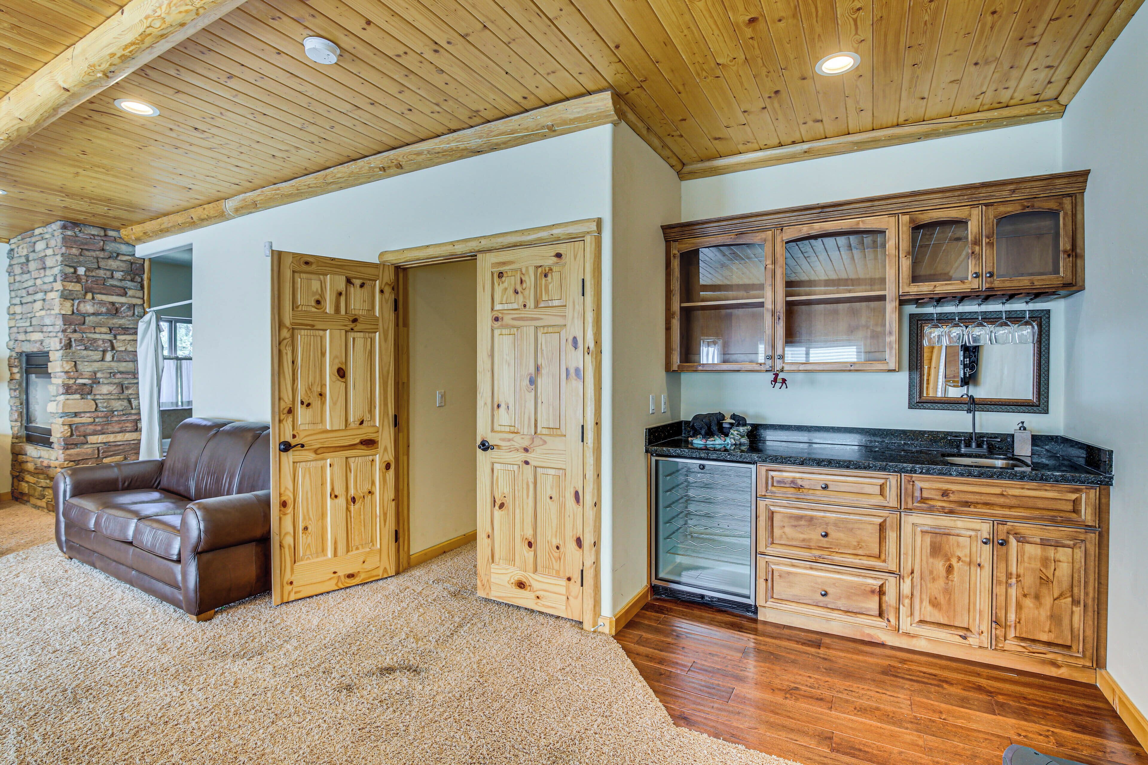 A spacious living area with a brown leather couch, wooden doors, a stone fireplace, and a kitchen with wooden cabinets and a black countertop.