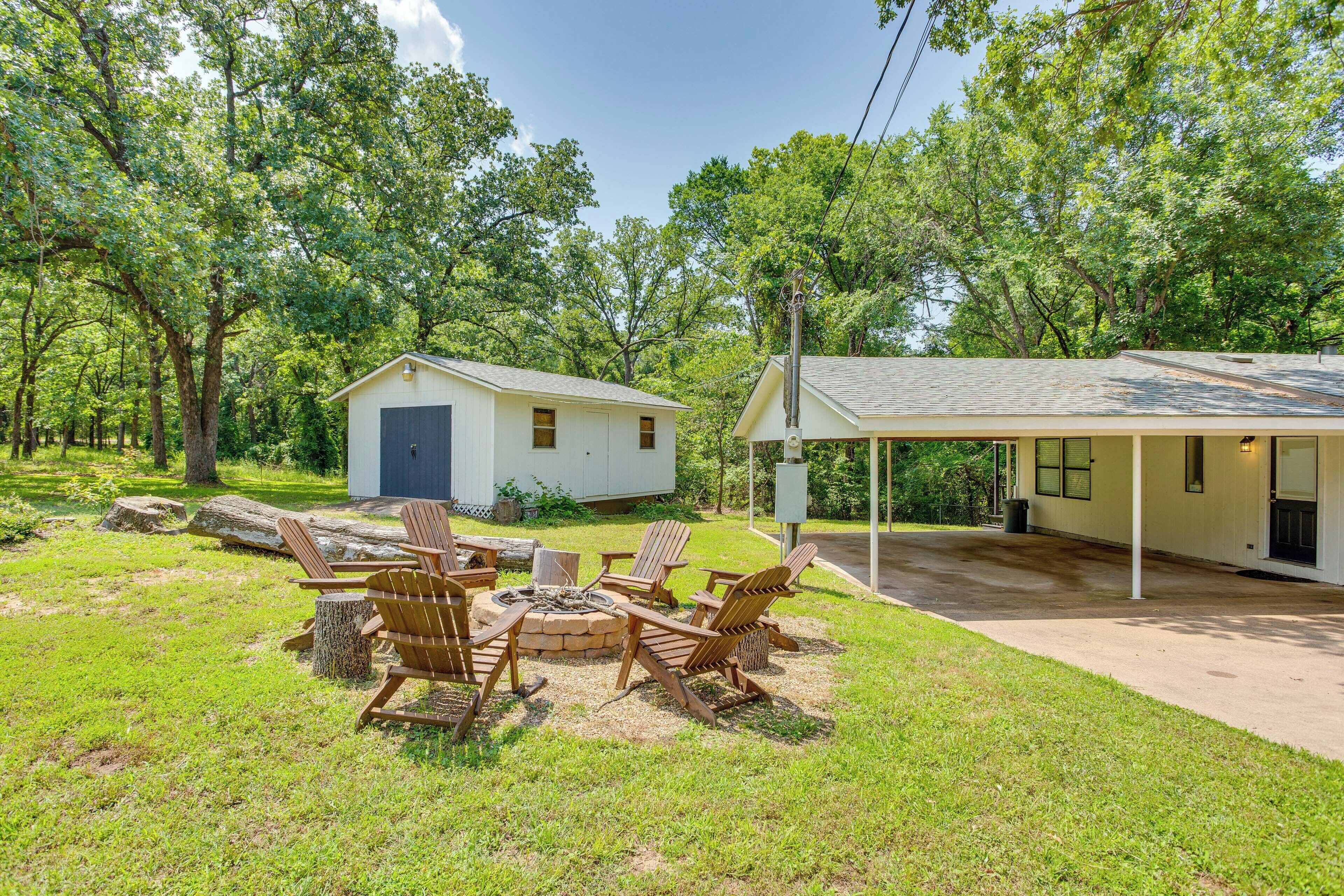 A covered outdoor seating area with Adirondack chairs and a fire pit, surrounded by a grassy yard and trees.