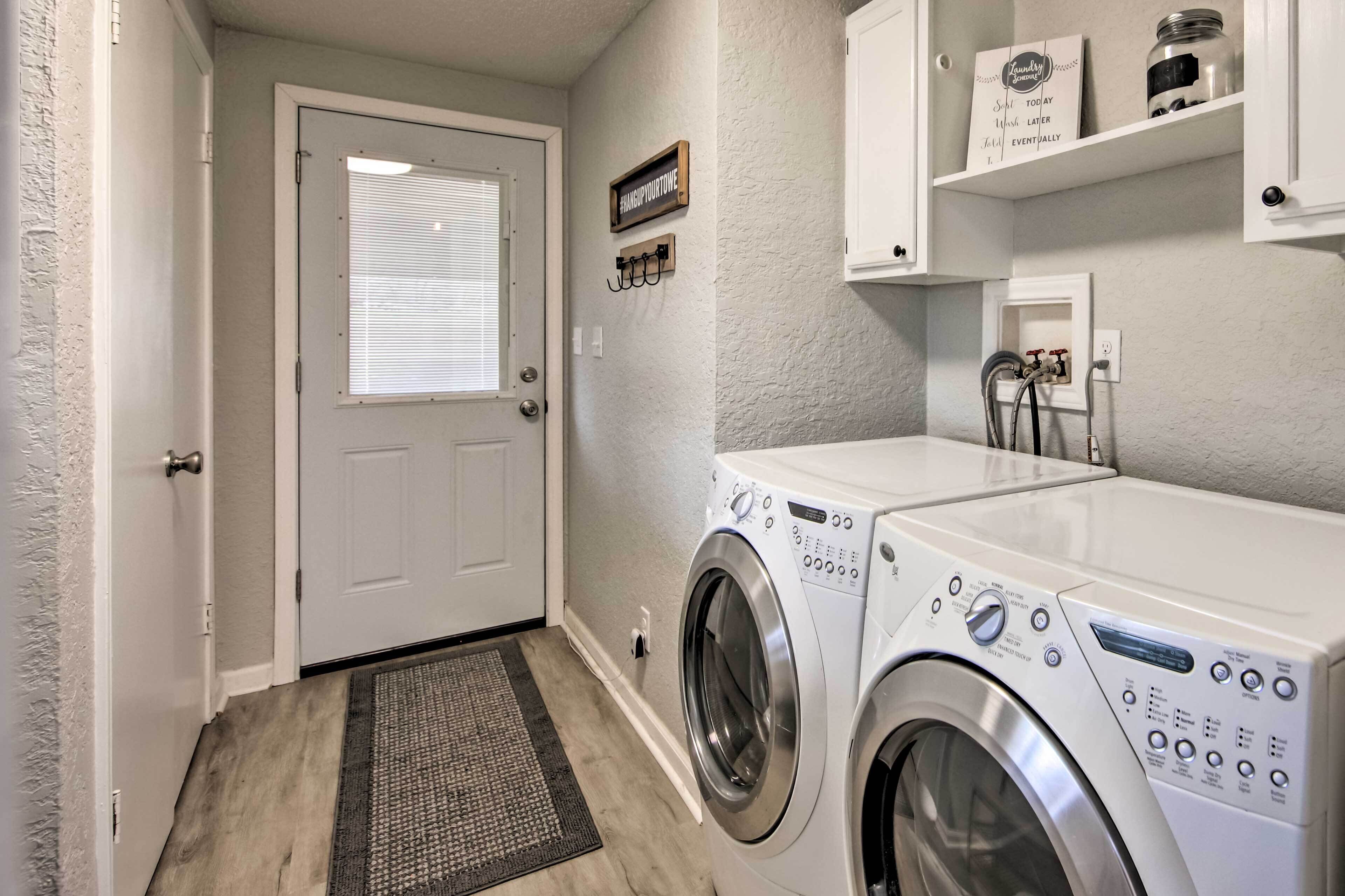 A laundry room with a front-loading washer and dryer, a door with a window, and a small shelf with jars and a sign.