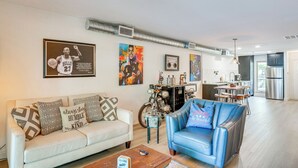 Uma sala de estar com um sofá de cor clara, uma poltrona azul e uma mesa de centro. Há quadros nas paredes, uma motocicleta e uma área de cozinha ao fundo.