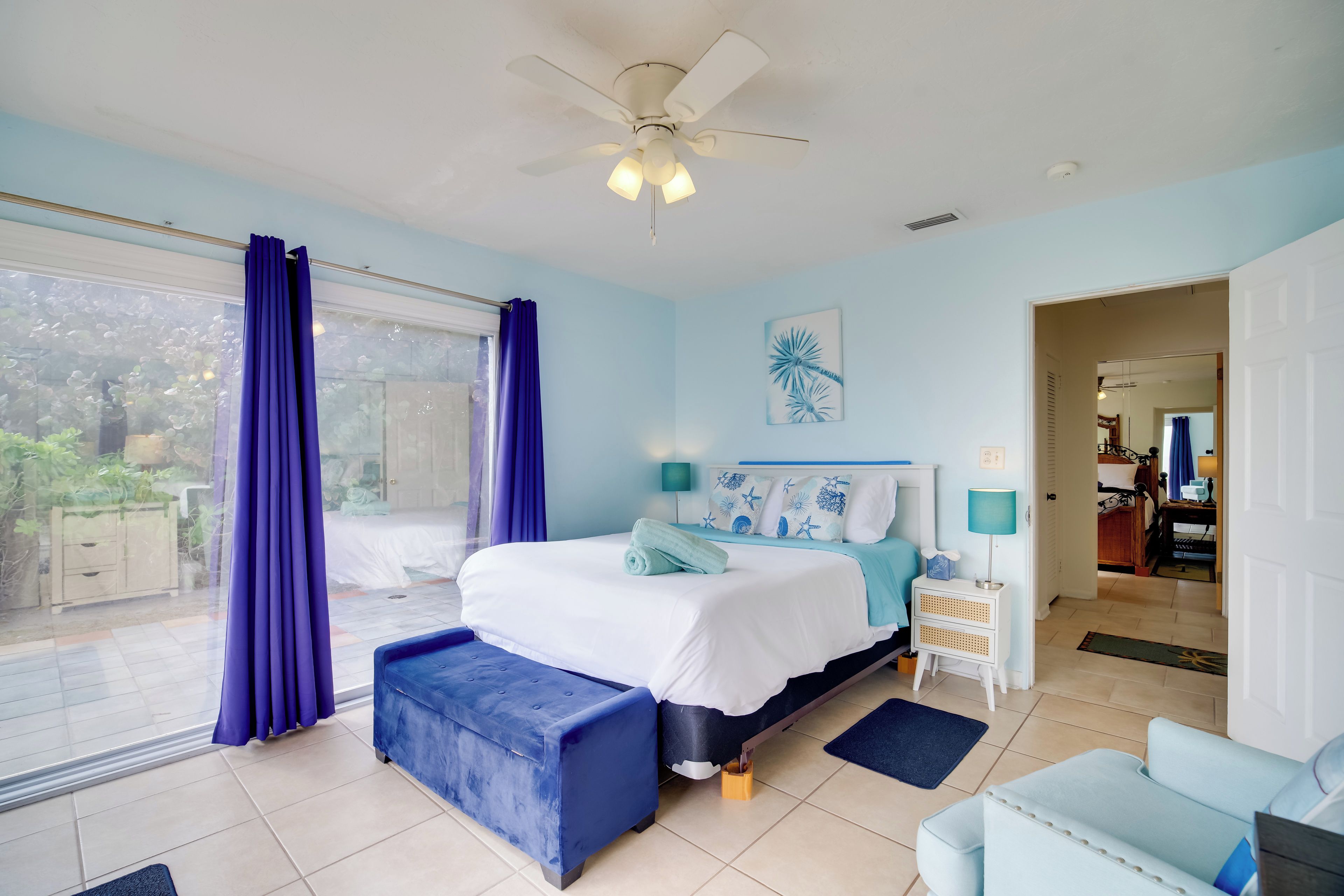 A bedroom with a bed, a blue bench, a ceiling fan, a window with curtains, a nightstand, and a door leading to another room.