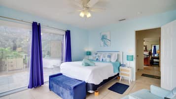 A bedroom with a bed, a blue bench, a ceiling fan, a window with curtains, a nightstand, and a door leading to another room.