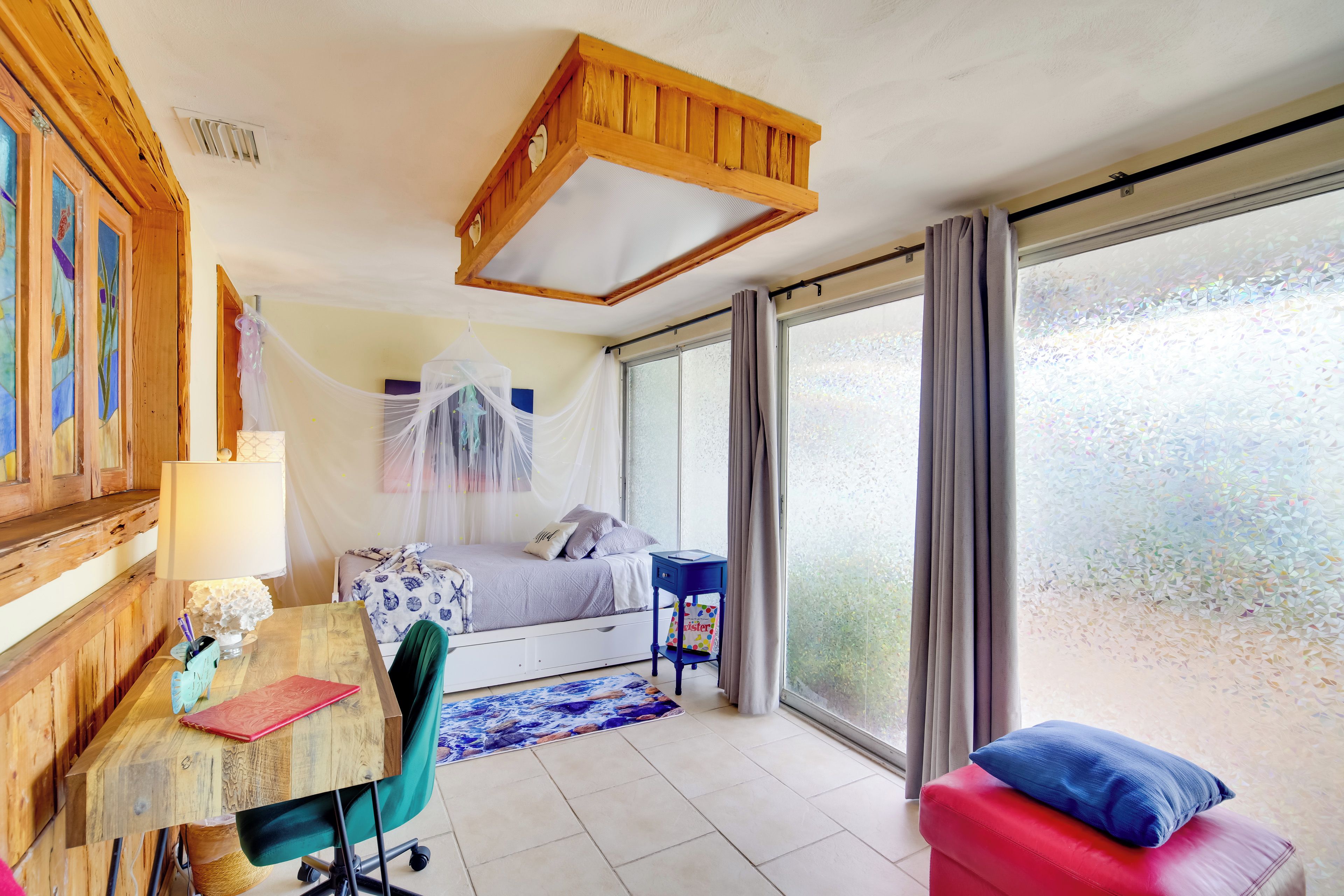 A bedroom with a bed, a desk, a chair, a window with curtains, and a wooden ceiling structure.