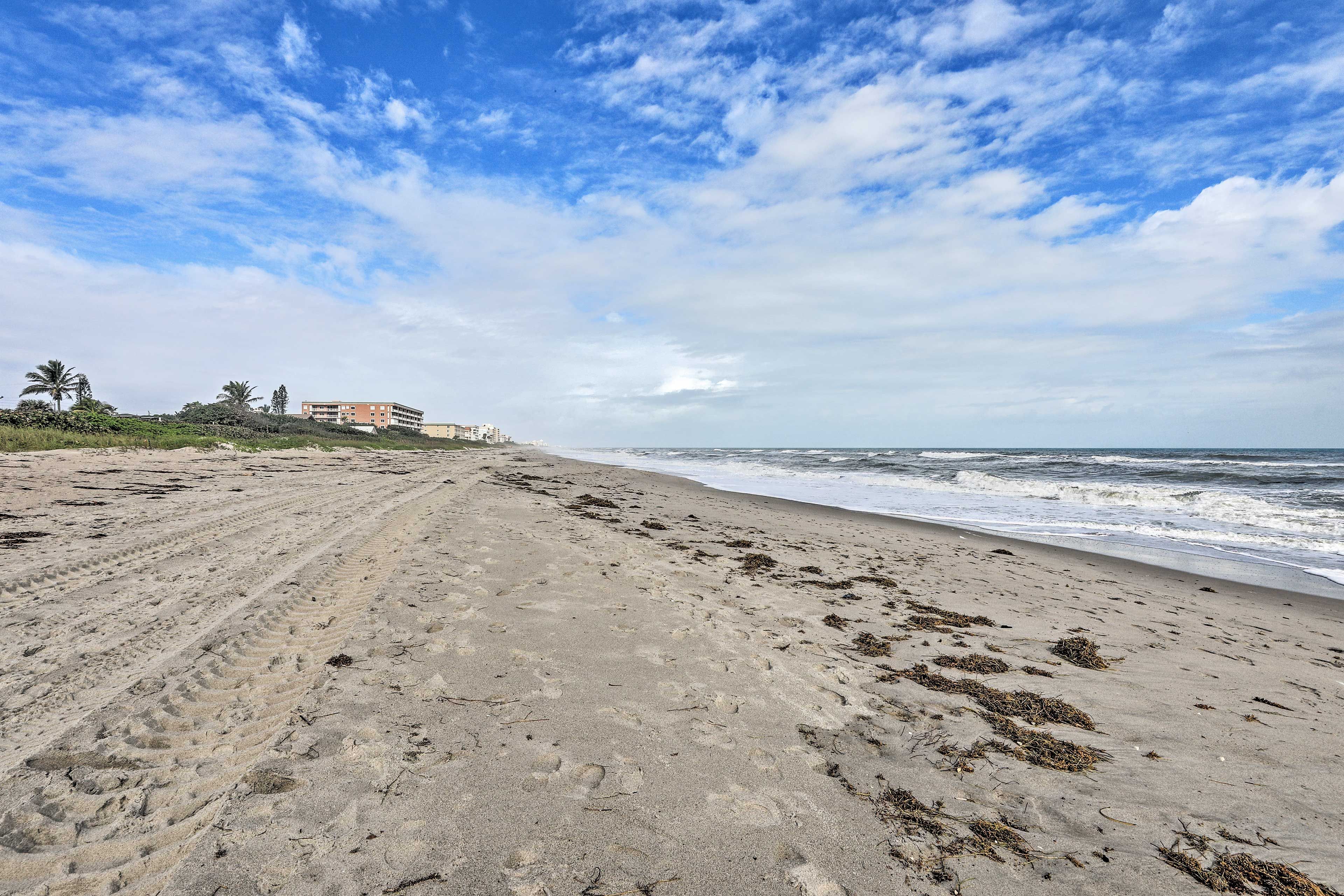 A beach with footprints, waves, and buildings in the background.